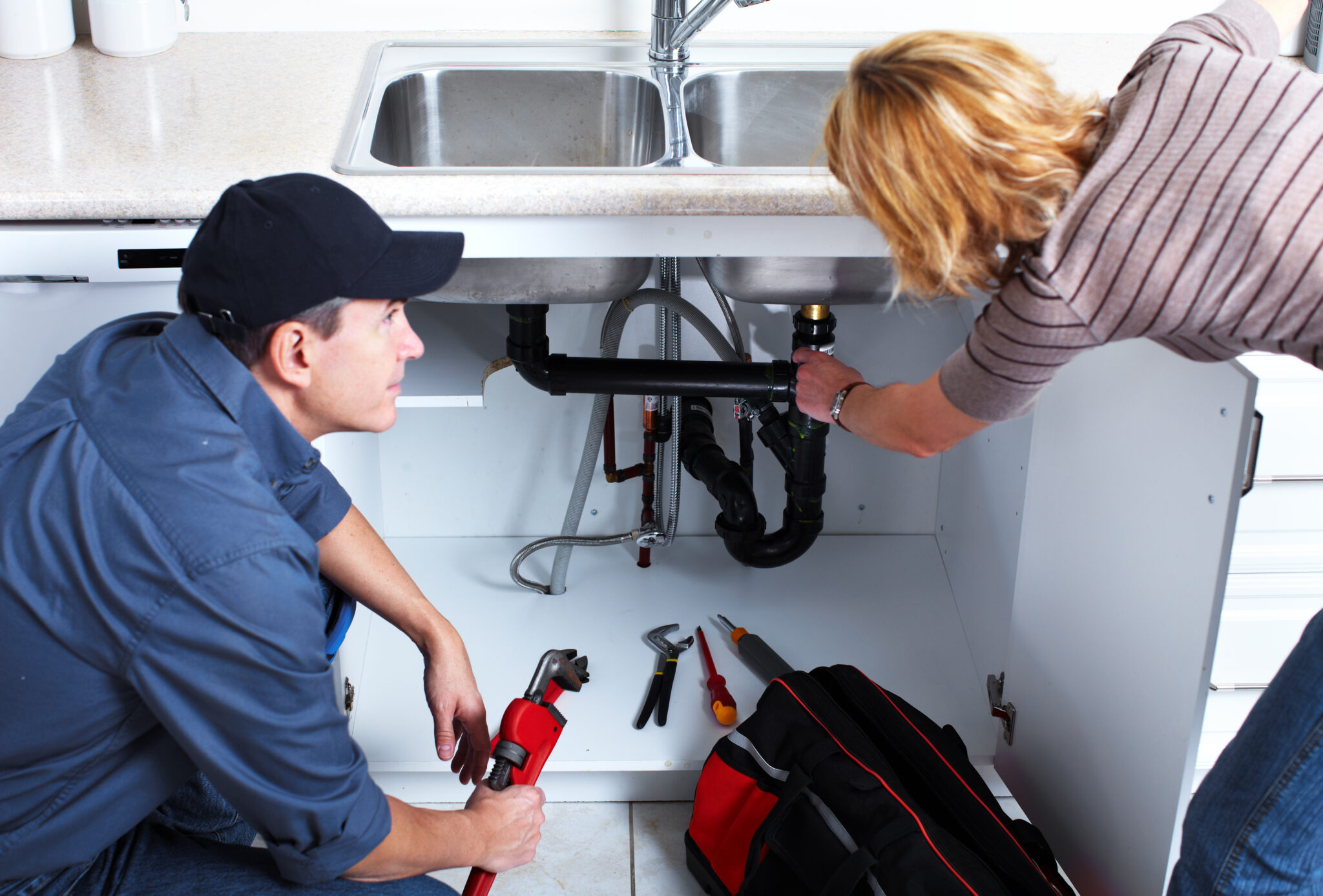 Plumber kneeling by an open kitchen cabinet explaining the pipework under a double sink to the homeowner